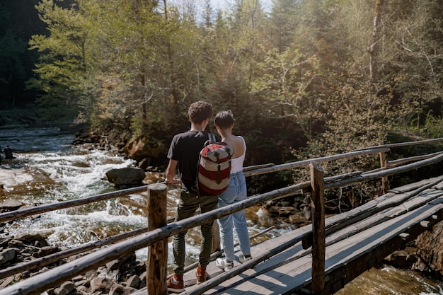 Vue arrière sur un couple de jeunes randonneurs debout sur un pont en bois au-dessus d'un ruisseau rapide et se serrant dans leurs bras