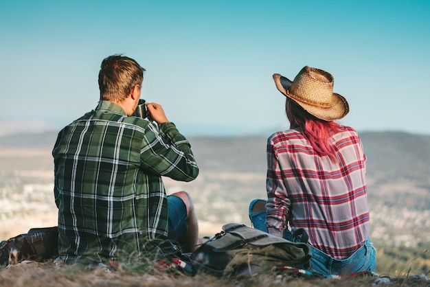 Vue Arrière D'un Couple De Jeunes Routards Heureux Se Reposant Après Avoir Atteint Le Sommet De La Montagne, Assis Sur Une Couverture, Buvant Du Thé Dans Une Bouteille Thermos Et Profitant De Beaux Paysages De Vue Sur La Vallée.