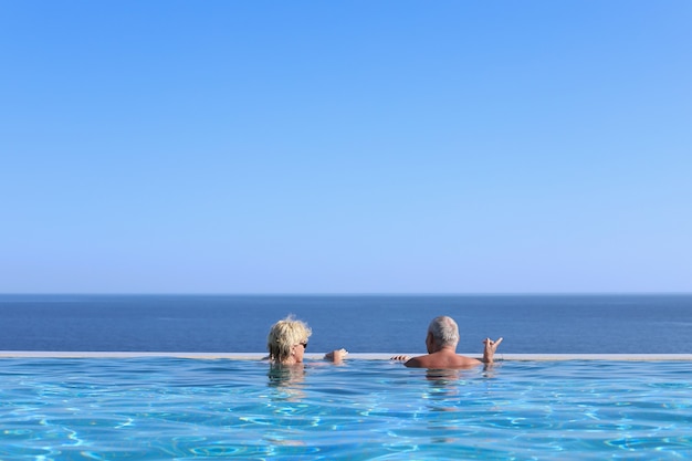 Vue arrière d'un couple de personnes âgées se reposant au bord d'une piscine bleue surplombant la mer