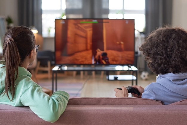 Vue arrière de deux adolescents assis sur un canapé devant un grand écran de télévision et jouant à un jeu vidéo à la maison