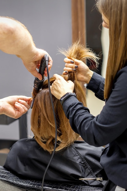 La vue arrière de deux coiffeurs frise les cheveux d'une jeune femme avec un fer à repasser électrique