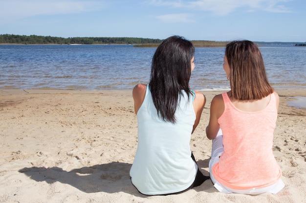 Vue arrière deux jeunes filles en forme s'asseoir sur la plage de sable regardent la côte de la mer