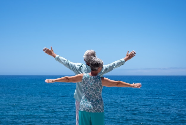 Vue arrière de deux personnes âgées debout à la mer avec les bras tendus, regardant l'horizon profitant de l'été et de la liberté