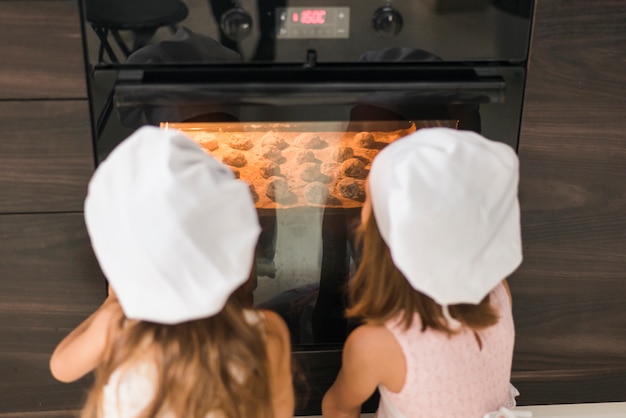 Photo vue arrière de deux soeurs en toque regardant un plateau à biscuits au four