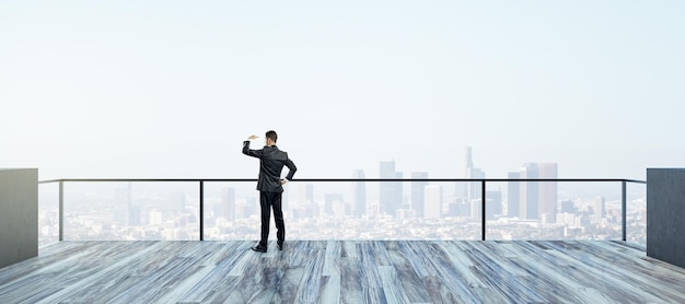 Vue arrière du jeune homme d'affaires regardant au loin sur le toit avec un ciel lumineux et une large vue sur la ville Succès futur et concept de vision