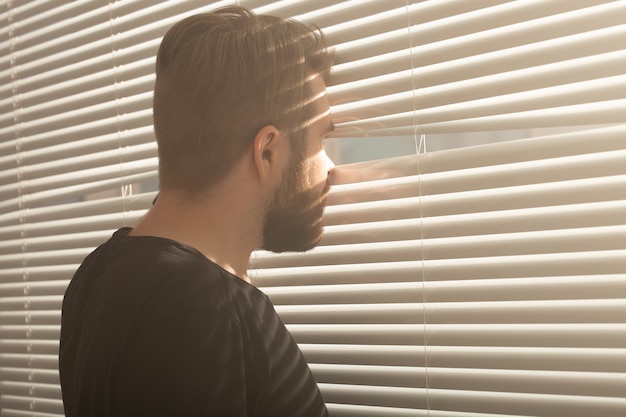 Vue arrière du jeune homme avec barbe jette un coup d'œil à travers le trou dans les stores