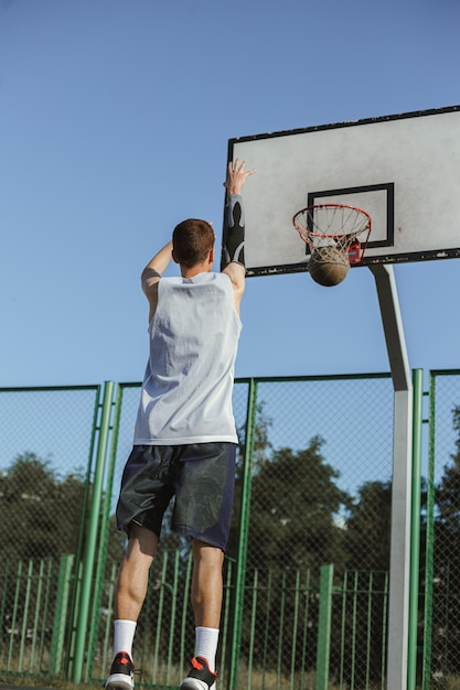 Vue arrière du jeune homme jouant au streetball et lançant la balle dans le cerceau sur le terrain de basket