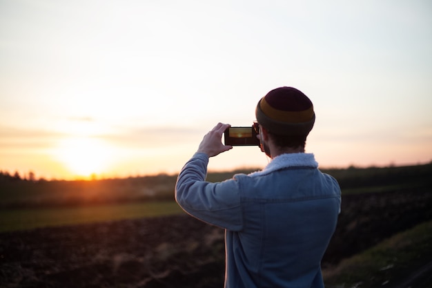 Vue arrière du jeune homme prendre des photos du coucher du soleil par smartphone