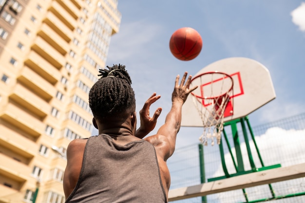 Photo vue arrière du jeune joueur de basket-ball africain lancer la balle dans le panier lors de la formation en milieu urbain