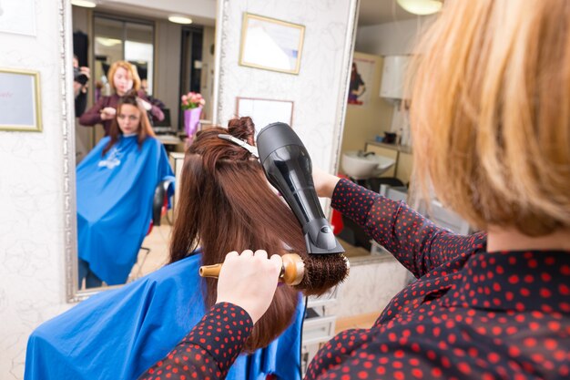 Photo vue arrière du jeune styliste blonde séchant les cheveux d'une cliente brune à l'aide d'un sèche-cheveux à main et d'une brosse ronde dans un salon avec une réflexion floue dans un grand miroir sur toute la longueur en arrière-plan