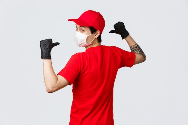 Vue arrière du livreur asiatique en masque médical et gants de protection, porter un bonnet rouge, un t-shirt, se tourner vers la caméra, pointant vers l'arrière pour montrer le logo de l'entreprise sur l'uniforme, la logistique, le courrier et le concept de magasinage