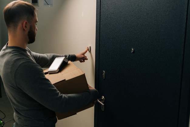 Vue arrière du livreur sonnant à la porte de l'appartement du client tenant une boîte en carton et un terminal sans fil POS de paiement sans contact