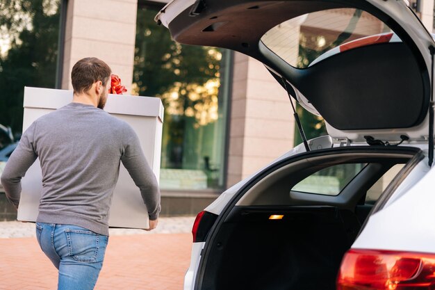 Vue arrière du livreur sort une grande boîte blanche festive avec un bel arc rouge du coffre de la voiture pour la livraison au client, à l'extérieur. Courrier se préparant à sortir le colis et à le livrer au client.