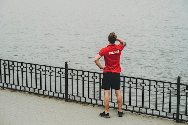 Vue arrière du mâle en chemise et short formateur rouge debout sur le remblai et à la distance à la surface de l'eau avec la main au front