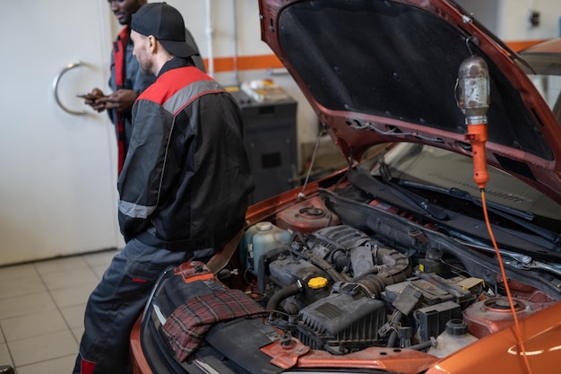 Vue arrière du mécanicien masculin prenant une pause dans un atelier automobile avec capot de voiture ouvert et moteur au premier plan, espace de copie