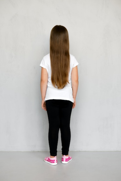 Vue arrière du mignon jeune enfant aux cheveux longs en t-shirt blanc et pantalon de survêtement noir posant