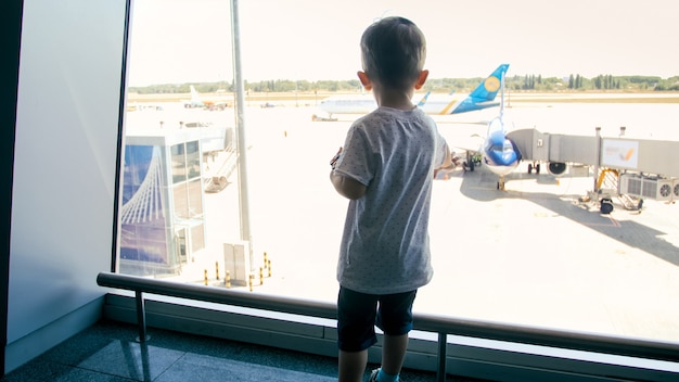 Vue arrière du mignon petit garçon regardant à travers la fenêtre du terminal de l'aéroport.