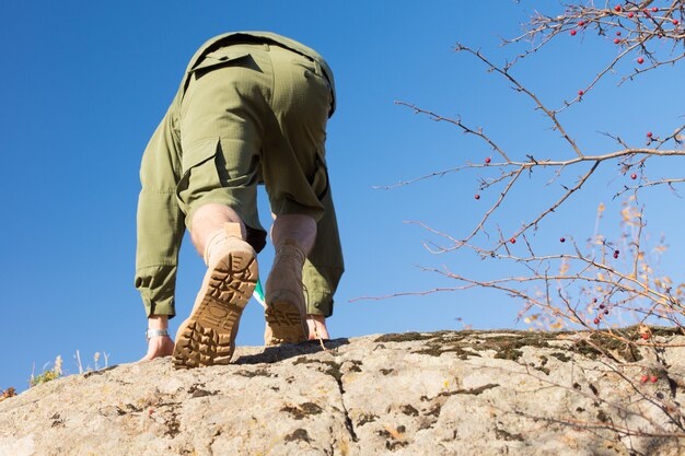 Vue arrière du scout blanc en uniforme escaladant un gros rocher sur un climat ensoleillé. Capturé avec fond de ciel bleu clair.