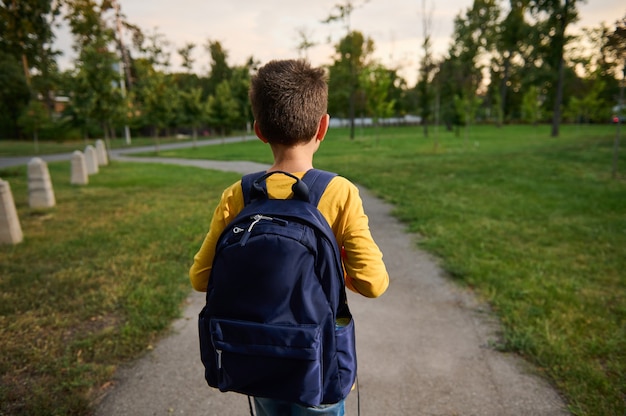 Vue arrière d'un écolier avec sac à dos cartable marchant sur le chemin dans un parc public, rentrant à la maison après l'école