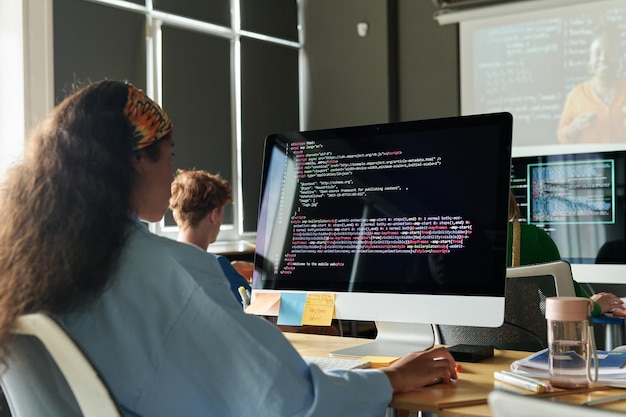Vue arrière d'une écolière assise au bureau devant un écran d'ordinateur et travaillant avec des codes informatiques