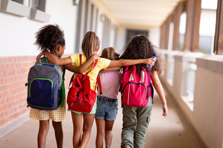 Photo vue arrière d'écolières élémentaires multiraciales avec des sacs à dos et des bras autour de la marche dans le couloir