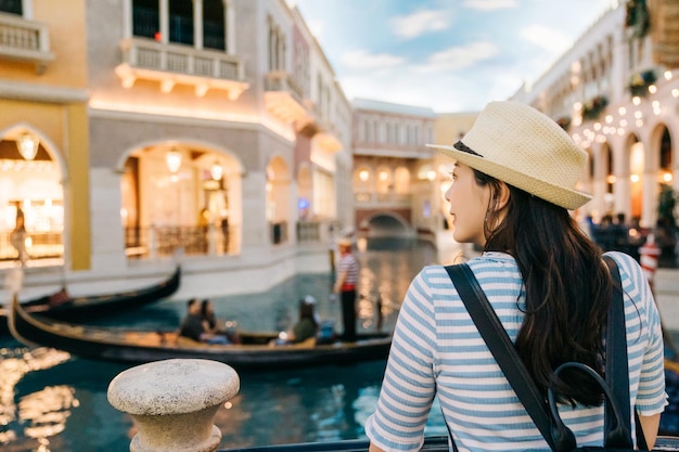 Photo vue arrière de l'élégante femme voyageuse avec chapeau de soleil profiter de la vue sur le canal en passant par la gondole dans le célèbre hôtel de las vegas. jeune fille backpacker souriante à la recherche d'un bateau pendant le coucher du soleil à l'extérieur lors d'un voyage d'été