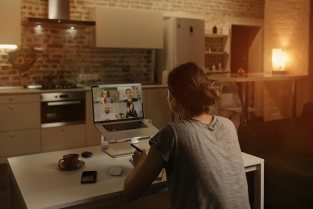 Vue arrière d'une employée qui travaille à distance et parle à ses collègues lors d'une vidéoconférence sur un ordinateur portable depuis la maison. Une équipe commerciale multiethnique lors d'une réunion en ligne le soir.