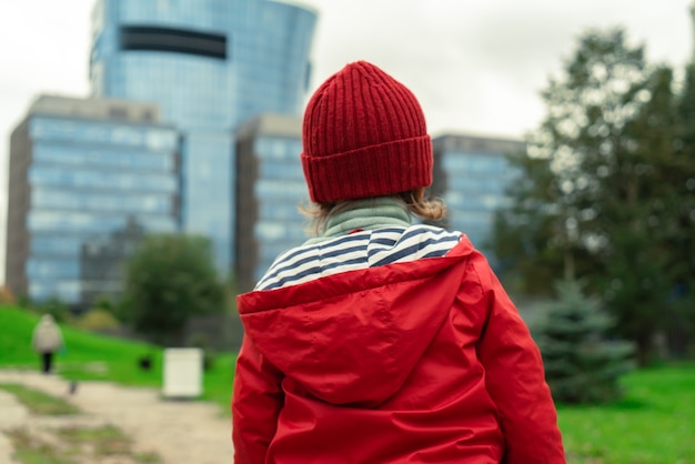 Vue arrière d'un enfant dans une veste et un chapeau rouges se tenant sur le fond d'une vue moderne de paysage urbain