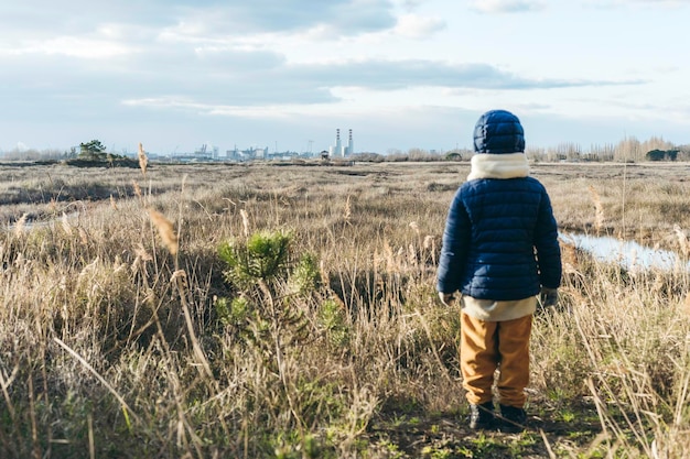 Vue arrière d'un enfant portant des vêtements d'hiver debout devant un paysage sauvage et des usines en arrière-plan Concept d'activisme et de protection de l'environnement Avenir de l'enfance pour les nouvelles générations