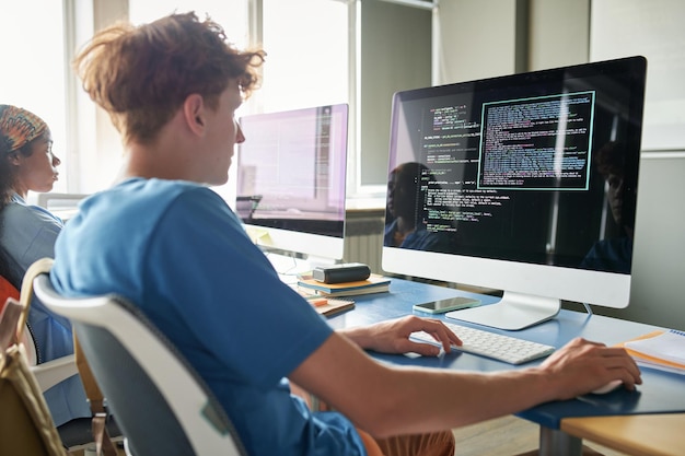 Vue arrière d'un étudiant assis au bureau avec un ordinateur et tapant des codes pendant un cours d'informatique