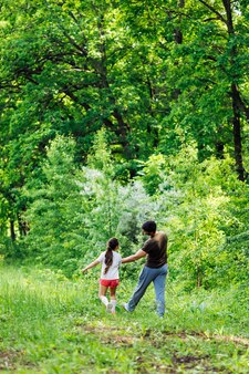 Vue arrière d'une famille heureuse marchant dans la forêt du parc autour d'arbres verts s'amusant petite fille tenant la main du père