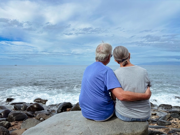 Vue arrière d'une famille liée, un couple de personnes âgées assis face à la mer sur la plage, s'embrassant et regardant l'horizon.