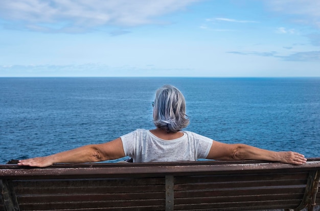 Vue arrière d'une femme âgée assise sur un banc en bois, profitant de la détente et de la nature. Mer bleue et ciel sur fond - moment positif pendant une retraite heureuse