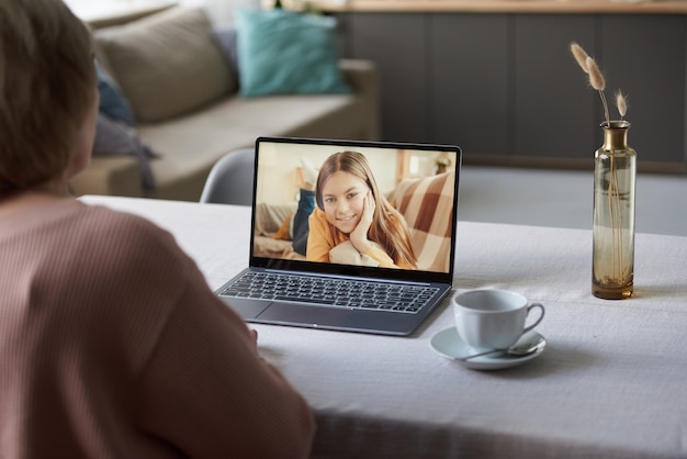 Vue arrière d'une femme âgée assise à la table devant un ordinateur portable et parlant à sa petite-fille en ligne à la maison