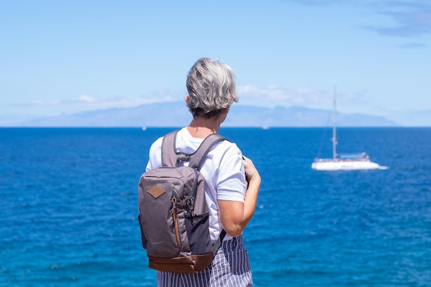 Vue arrière d'une femme âgée avec sac à dos debout et regardant la mer et l'île à l'horizon vacances d'été ou retraite