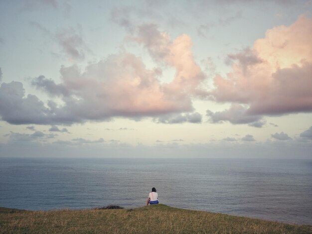 Vue arrière d'une femme assise sur une montagne par la mer contre un ciel nuageux