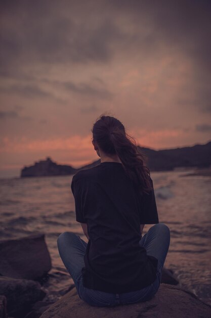 Vue arrière d'une femme assise sur un rocher sur la plage au coucher du soleil