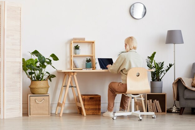 Vue arrière de la femme assise à la table et travaillant sur un ordinateur portable dans la chambre à la maison