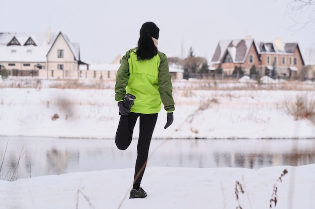 Vue arrière de la femme brune faisant de l'exercice quad stretch tout en étirant la jambe à la côte de la rivière d'hiver