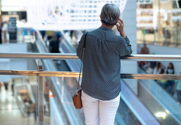 Vue arrière d'une femme caucasienne âgée debout à l'intérieur d'un centre commercial regardant la foule et l'escalator