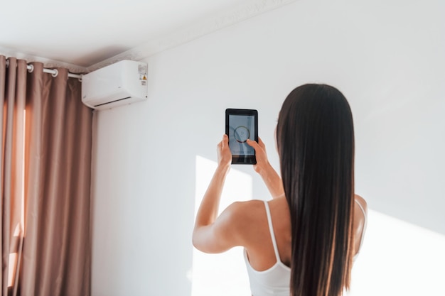 Vue arrière d'une femme debout à l'intérieur avec un téléphone et contrôlant la technologie de la maison intelligente