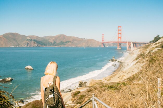 Photo vue arrière d'une femme debout sur le rivage de la mer par le pont de la porte d'or contre un ciel bleu clair