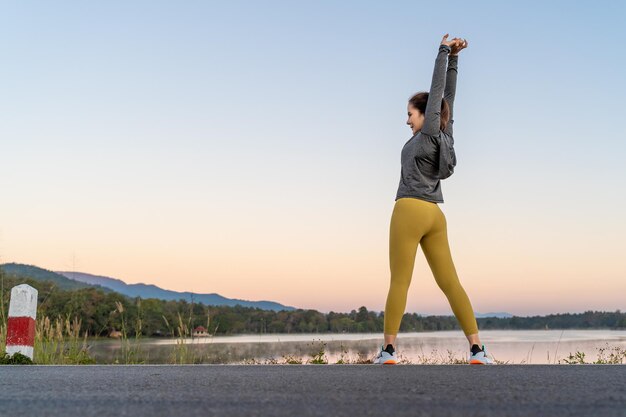 Vue arrière d'une femme étirant ses bras et ses jambes avant son exercice tôt le matin dans un parc de lac local avec les montagnes du ciel du lever du soleil du matin et la salle de fond du lac pour l'espace de copie