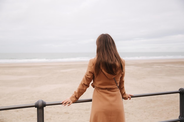 Vue arrière de la femme en manteau brun debout le long de la mer du Nord et regardant les vagues
