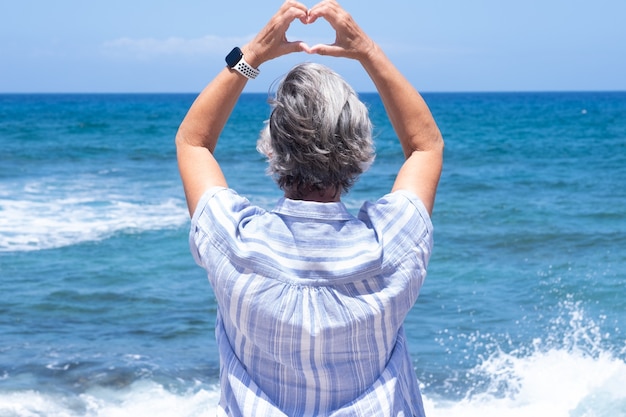 Vue arrière d'une femme mature avec un chapeau bleu regardant l'horizon au-dessus de l'eau en forme de coeur avec les mains. Concept d'amour et de liberté