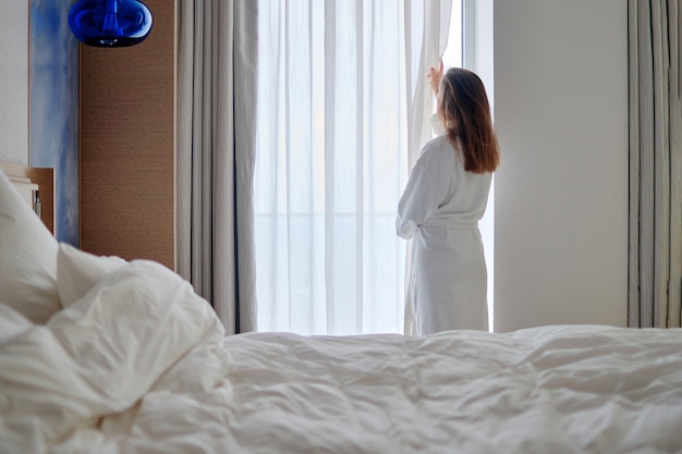 Vue arrière de la femme portant un peignoir se réveiller, ouvrir les rideaux et profiter du bonjour tout en vous relaxant dans une chambre confortable dans la chambre d'hôtel.