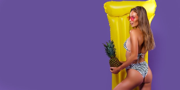 Vue arrière d'une femme qui pose en bikini avec un matelas gonflable jaune tout en tenant un ananas