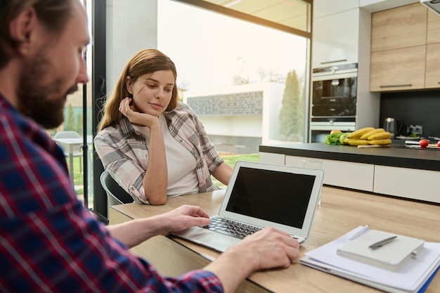 Vue arrière d'une femme regardant l'écran de l'ordinateur portable de son mari travaillant à distance depuis la maison Pile de documents et bloc-notes allongé sur le bureau Copier l'espace pour le texte