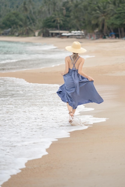 Vue arrière de la femme en robe bleue et chapeau de paille à pied sur la plage