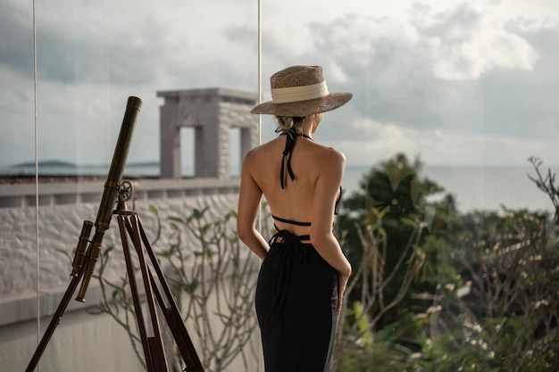 Vue arrière d'une femme en robe noire debout près du télescope sur un balcon regardant la vue sur l'océan.